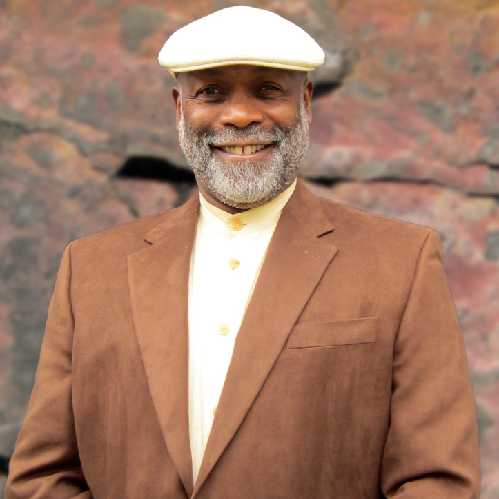 Image of artist Lawrence Diggs. Diggs is shown from the waist up, wearing a brown suit jacket, off-shite collared shirt, and white cap. He is smiling through a full grey beard. 