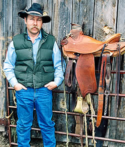 Robert Dennis (man) stands at left of handmade leather saddle on metal fence