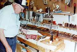 Man (Stan Fillingsness) stands at workbench, looks through book. Wood working tools line back of bench.