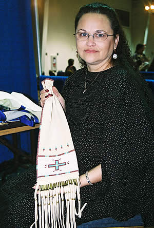 Woman (Linda Thompson) stands, presenting a beaded pipe bag. Fringe at bottom of the bag. 