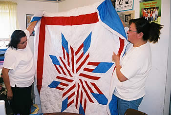 Two women (Renee Turning Heart at left and Patricia Kennedy) stand holding a red, white, and blue star quilt.