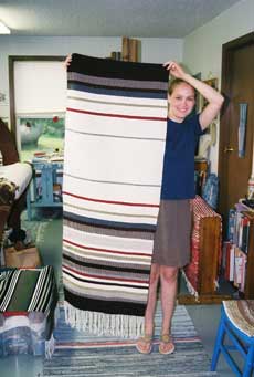 Woman (Marianne Klipfel) standing with woven piece at left, as tall as she is. Piece has white, tan, black, and red horizontal stripes and lond fringe at bottom (facing floor).