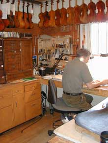 John Waddle seated at work bench working in violin repair shop. Violins hang over his head. 