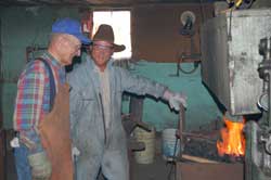 Tro men standing in front of forge (open flame) with metal tools in shop.