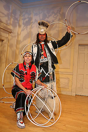 Jasmine Pickner (woman kneeling at left of) and Dallas Chief Eagle (man standing at right and behind woman) together in red and black traditional clothing with hoops.