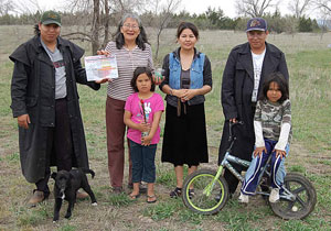 Four adults (two men at ends and two women in middle) stand in a row behind two youth (one on a bicycle) and small dog in open field. 