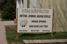 Yarn shop sign, reads "Natural colored wool studio. Knitting, spinning, weaving supplies. Unique apparel. Home decor and gift items."