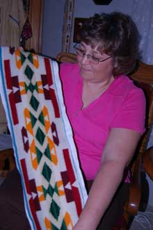 Tamara Fonder (woman) shows beadwork on a Pendleton blanket (long banner with red, dark red, orange, and green patterns)