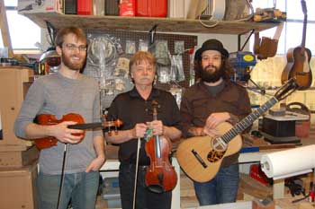 Three men standing in a row with instruments in a small shop.