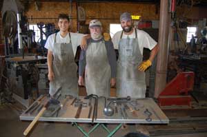 Three men standing behind low table with steel items the men made together. Master blacksmith Jack Parks (center) and apprentices.