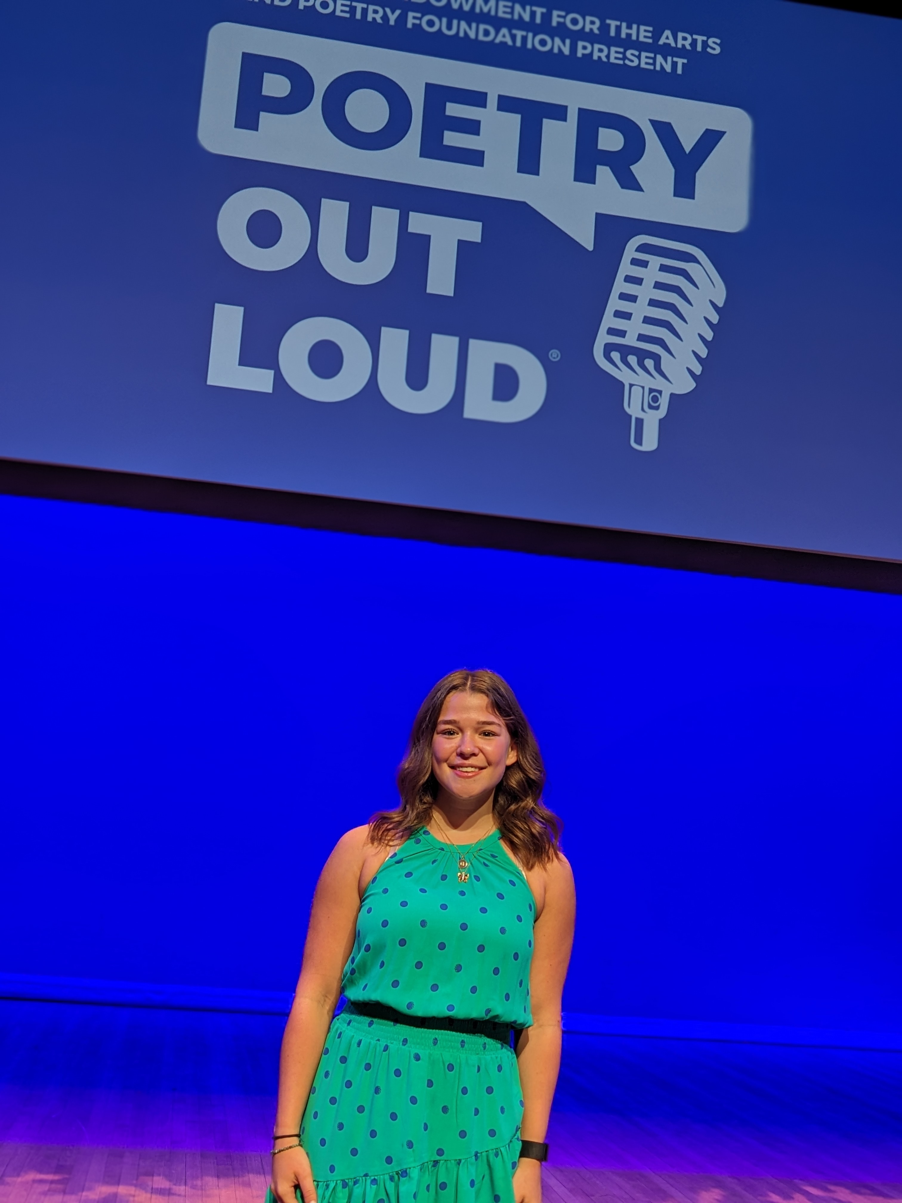 A young woman in a turquoise polka-dot dress stands on a stage, smiling at the camera. Behind her, a screen displays the "Poetry Out Loud" logo with a speech bubble and microphone icon.