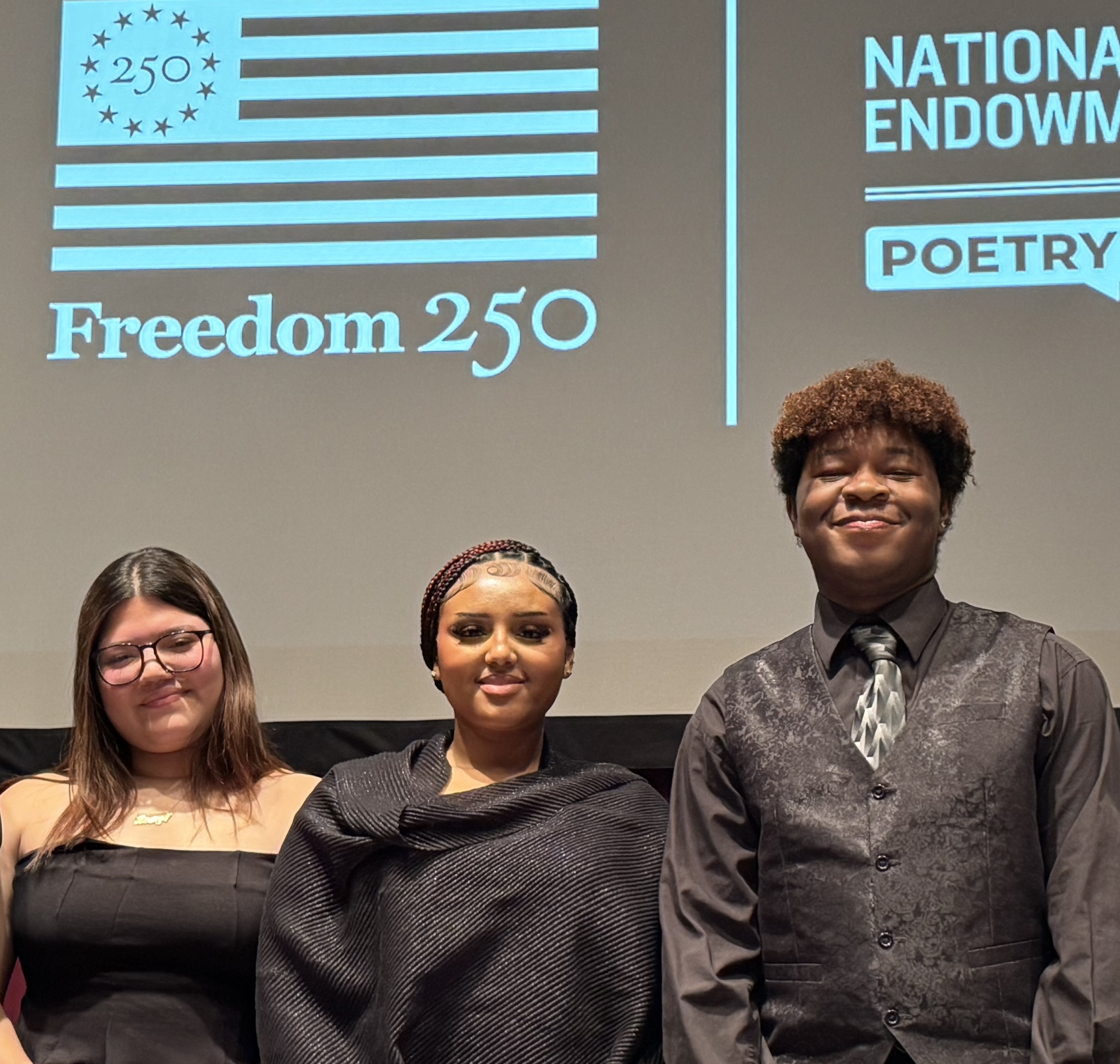Three young people stand on a stage in front of a projected screen that reads “Freedom 250” with a stylized American flag and poetry event signage.