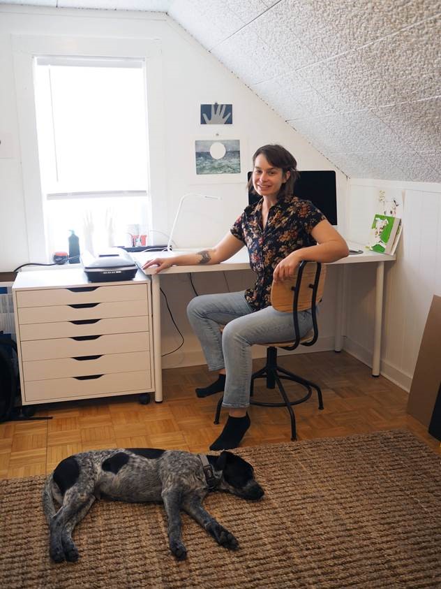Woman (Amber Zora)  seated at desk with dog laying on floor in front