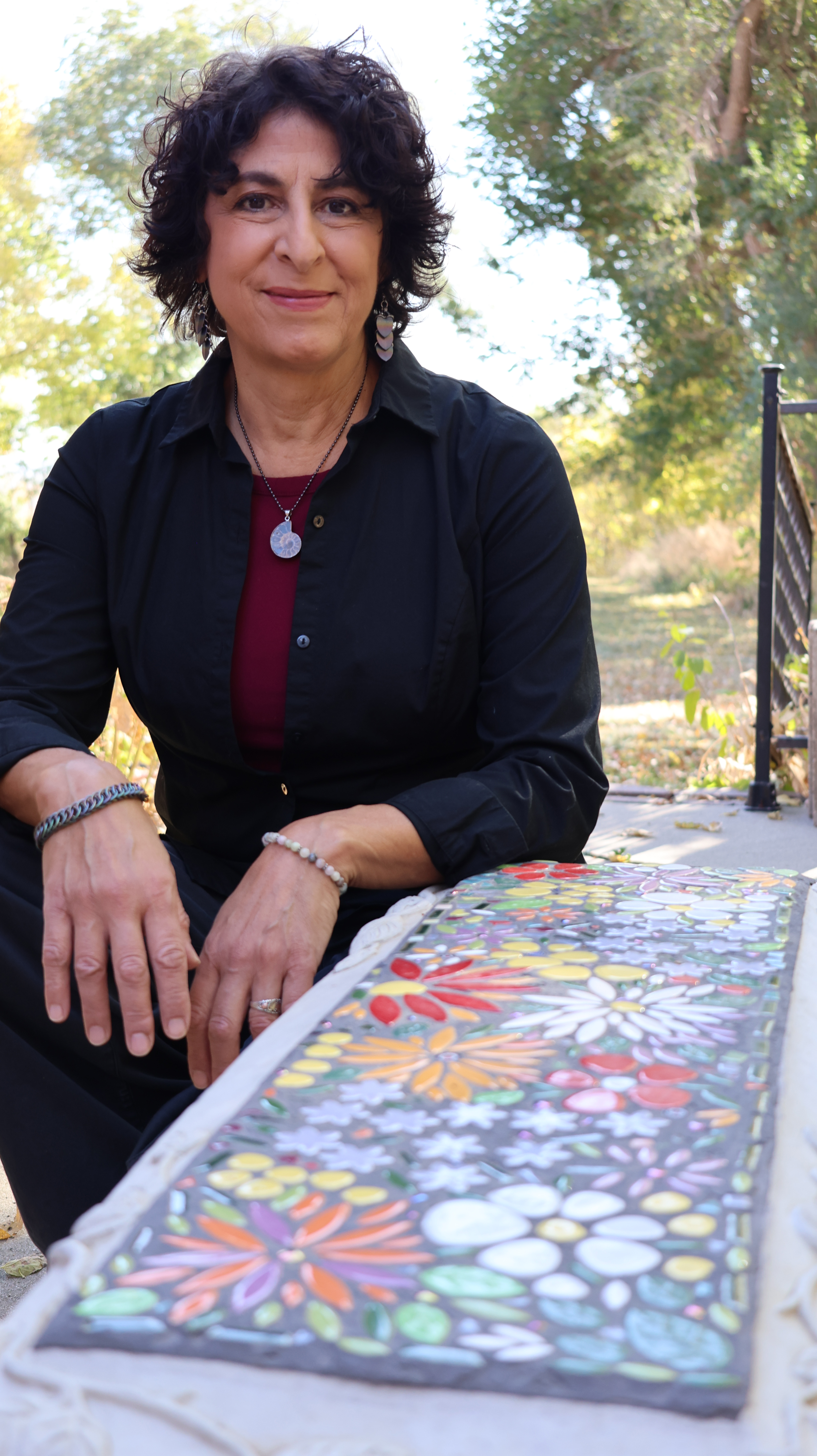 A person wearing a black shirt and red top, with a necklace and bracelets, sits outdoors in front of a colorful mosaic artwork featuring floral patterns.