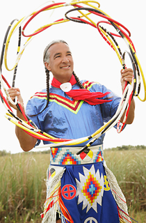 Man (Kevin Locke) stands with hoop. Dressed in traditional clothing.