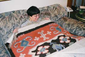 Woman (Diane Fields) sits on couch with weaving covering all but shoulders and head. Weaving is multicolored.